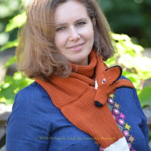 May include: A woman wearing a blue shirt with embroidered flowers and a knitted orange fox scarf. The scarf has a white tip and black eyes. The text "2015 Vitalina Craft by Tatsiana Matsyuk" is visible on the scarf.
