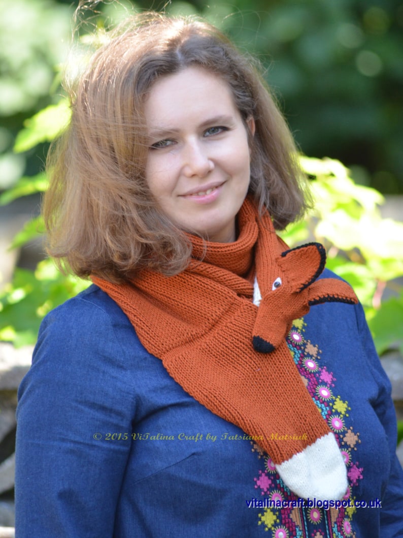 May include: A woman wearing a blue shirt with embroidered flowers and a knitted orange fox scarf. The scarf has a white tip and black eyes. The text "2015 Vitalina Craft by Tatsiana Matsyuk" is visible on the scarf.