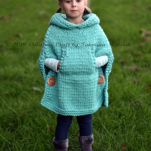 May include: A turquoise knitted poncho with a hood and two pockets. The poncho has wooden buttons on the pockets. A young girl is wearing the poncho and standing in a grassy field.