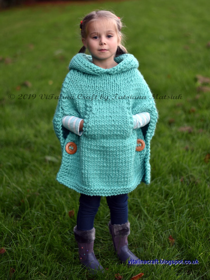 May include: A turquoise knitted poncho with a hood and two pockets. The poncho has wooden buttons on the pockets. A young girl is wearing the poncho and standing in a grassy field.