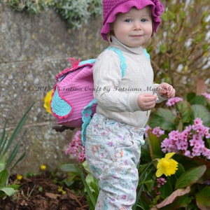 May include: A young child wearing a pink crocheted hat with a flower detail and a white crocheted backpack with a pink and blue owl design. The child is standing in a garden with pink and yellow flowers.