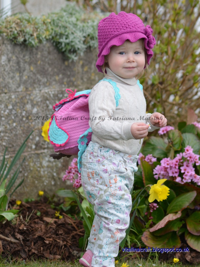 May include: A young child wearing a pink crocheted hat with a flower detail and a white crocheted backpack with a pink and blue owl design. The child is standing in a garden with pink and yellow flowers.