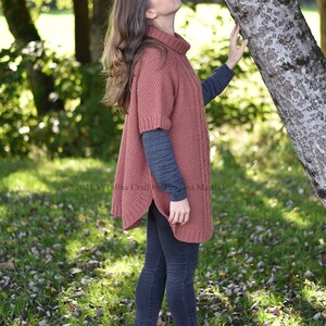 K&ouml;nnte beinhalten: Eine Frau tr&auml;gt einen rostigen Strickponcho mit Rollkragen, eine dunkle Jeans und rote Schuhe. Sie steht in einem grasbewachsenen Bereich mit heruntergefallenen Bl&auml;ttern und einem Baumstamm hinter ihr.