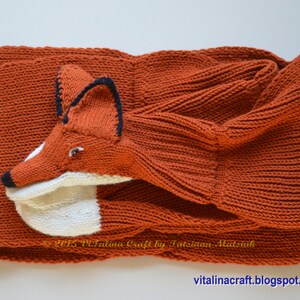 May include: A knitted orange fox scarf with a white face and black eyes. The scarf is folded in half and the fox's head is sticking out.