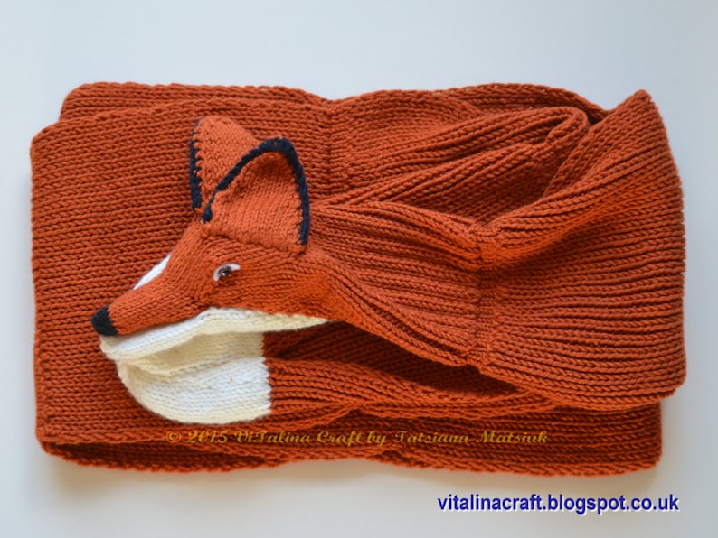 May include: A knitted orange fox scarf with a white face and black eyes. The scarf is folded in half and the fox's head is sticking out.