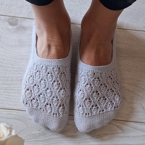 May include: Light grey knitted slipper socks with a detailed openwork design. The socks are worn on feet, with the ankles and lower legs visible. The socks are on a light wood floor.