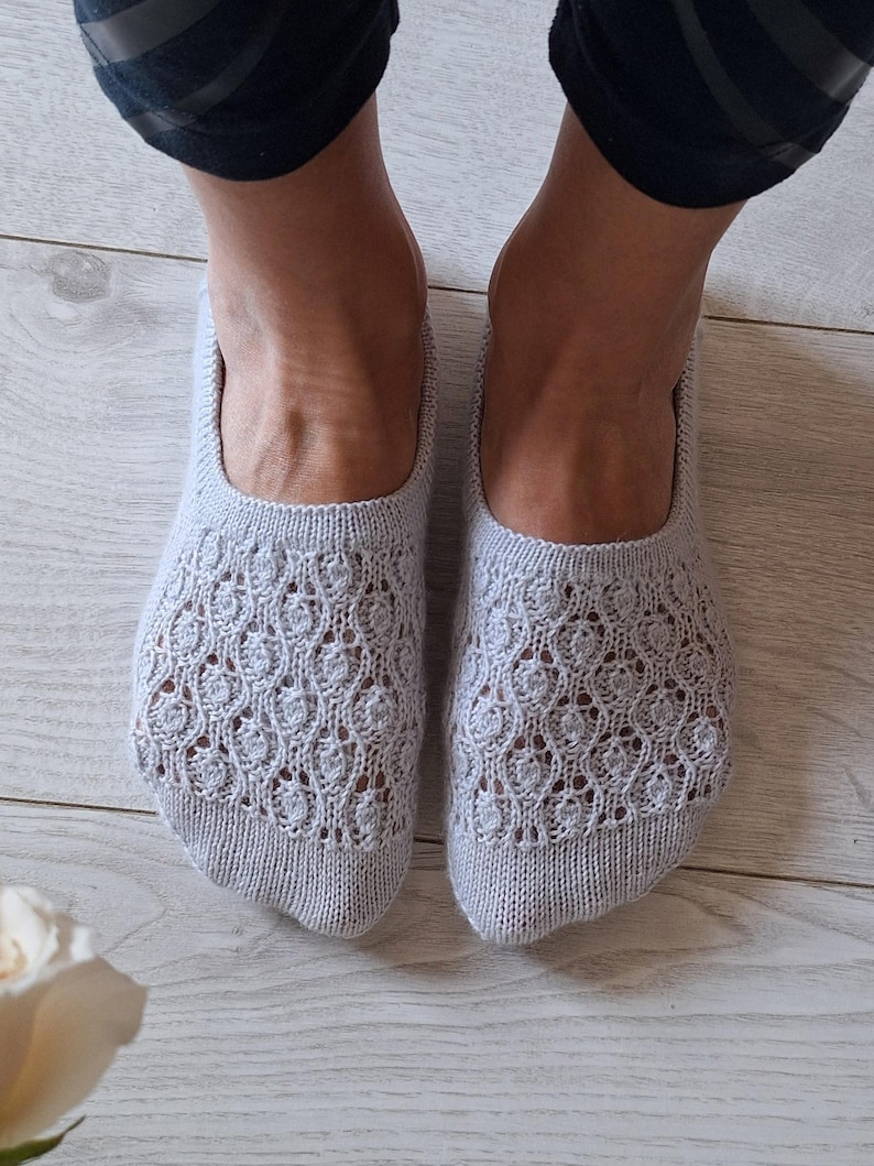 May include: Light grey knitted slipper socks with a detailed openwork design. The socks are worn on feet, with the ankles and lower legs visible. The socks are on a light wood floor.