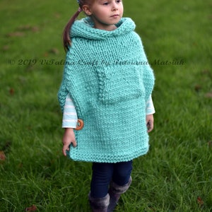 May include: A young girl wearing a turquoise knitted poncho with a wooden button. The poncho has a large hood and is made with a chunky yarn. The girl is standing in a grassy field.