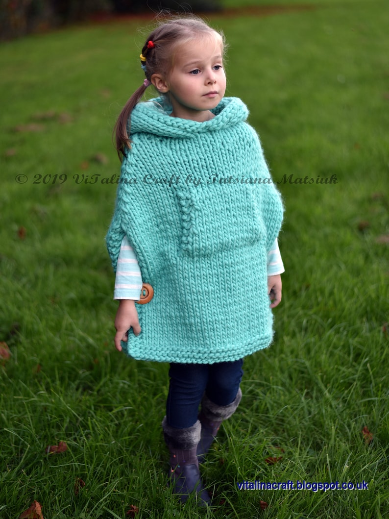 May include: A young girl wearing a turquoise knitted poncho with a wooden button. The poncho has a large hood and is made with a chunky yarn. The girl is standing in a grassy field.