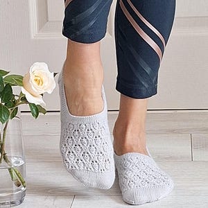 May include: Pale grey knitted socks with an openwork pattern. The socks are worn with dark blue leggings with rose gold stripes. A white rose in a glass vase is in the background.