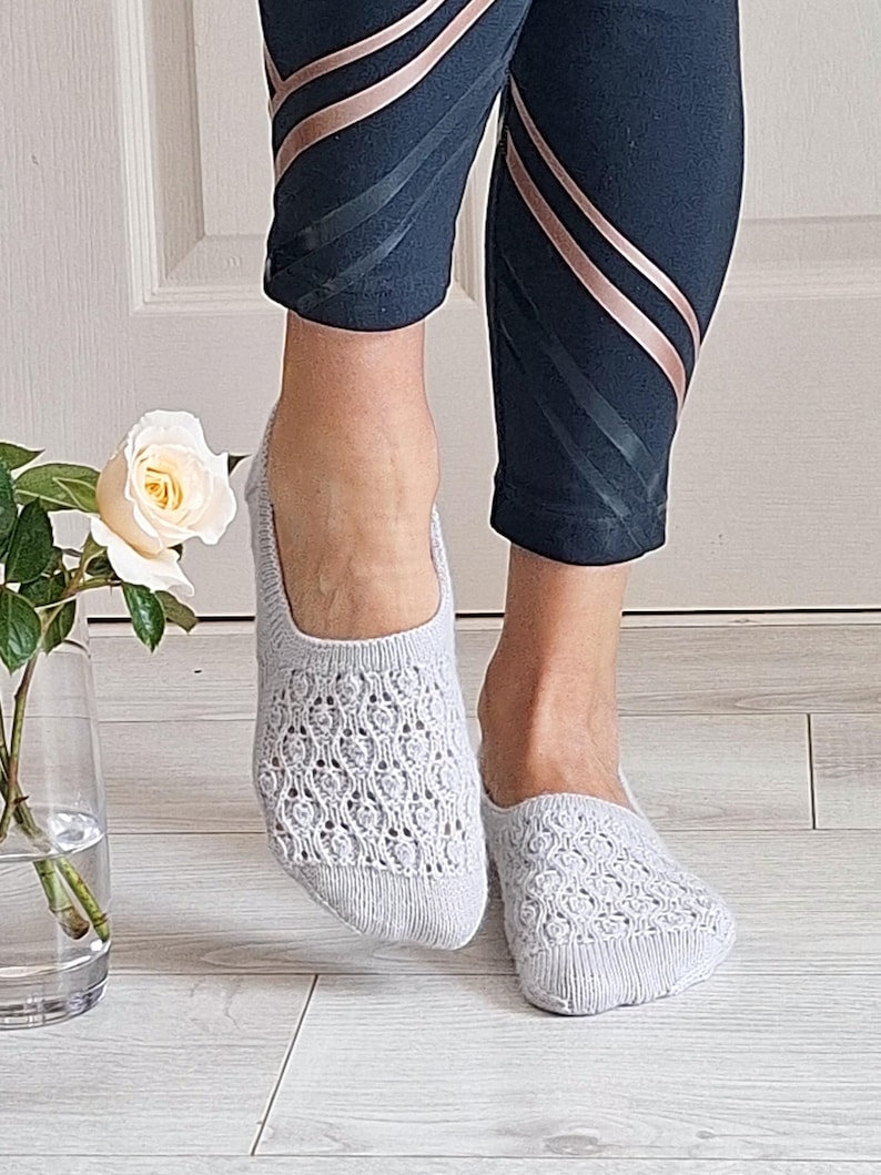 May include: Pale grey knitted socks with an openwork pattern. The socks are worn with dark blue leggings with rose gold stripes. A white rose in a glass vase is in the background.