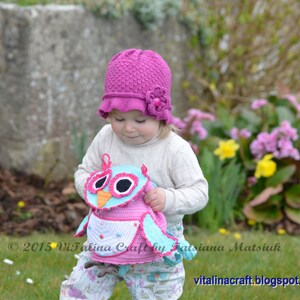 May include: A young child wearing a pink crocheted hat with a flower detail holds a crocheted owl toy. The owl is pink, blue, and white with black eyes. The child is standing in a grassy area with flowers in the background.