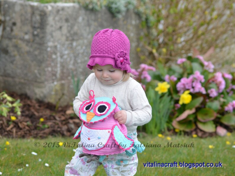 May include: A young child wearing a pink crocheted hat with a flower detail holds a crocheted owl toy. The owl is pink, blue, and white with black eyes. The child is standing in a grassy area with flowers in the background.