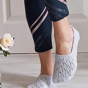 May include: Light grey open-weave socks with a floral design. The socks are worn with black leggings with rose gold and black stripes. A white rose is visible in the background.