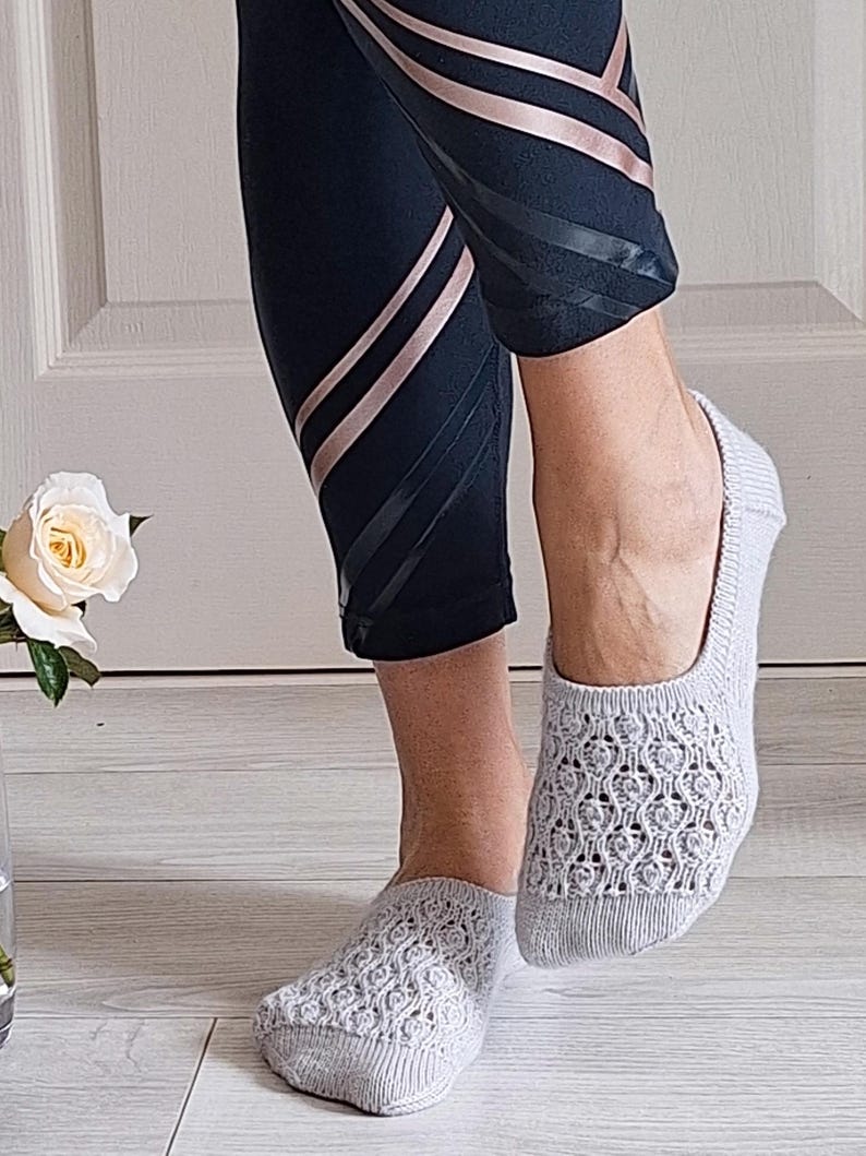 May include: Light grey open-weave socks with a floral design. The socks are worn with black leggings with rose gold and black stripes. A white rose is visible in the background.