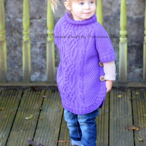 May include: A young girl wearing a purple knitted sweater with a cable knit pattern and blue jeans. The sweater has a turtleneck and a button on the side. The girl is standing on a wooden deck with fallen leaves.