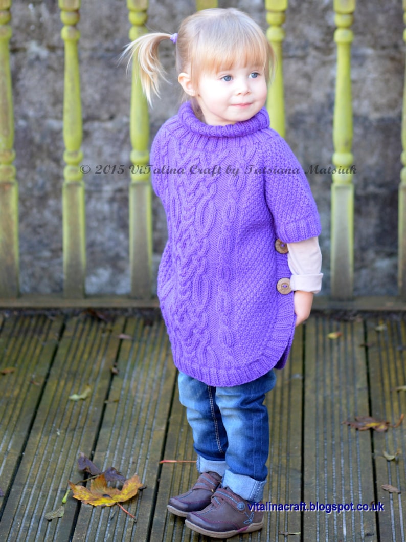 May include: A young girl wearing a purple knitted sweater with a cable knit pattern and blue jeans. The sweater has a turtleneck and a button on the side. The girl is standing on a wooden deck with fallen leaves.