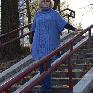 K&ouml;nnte beinhalten: Eine Frau tr&auml;gt einen blauen Strickponcho mit Rollkragen und einem Knopfdetail. Sie steht auf einer Treppe mit einem Gel&auml;nder.