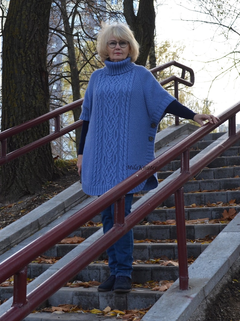 K&ouml;nnte beinhalten: Eine Frau tr&auml;gt einen blauen Strickponcho mit Rollkragen und einem Knopfdetail. Sie steht auf einer Treppe mit einem Gel&auml;nder.