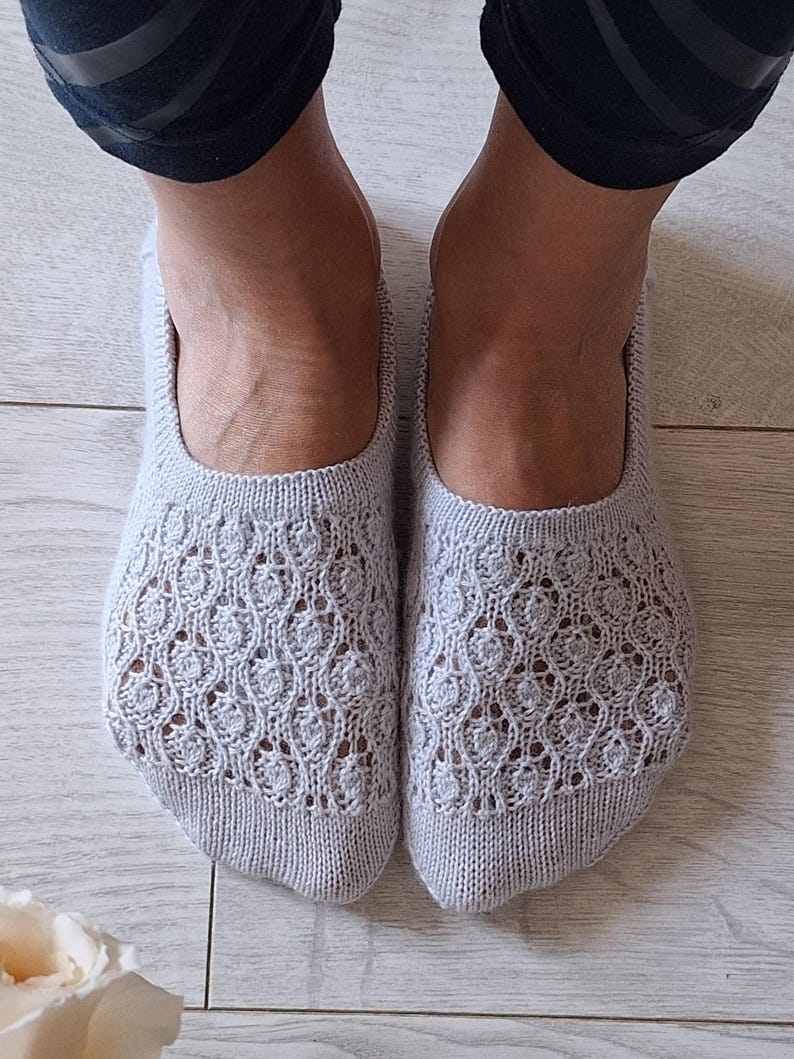 May include: Light grey knitted slipper socks with an openwork pattern. The socks are visible from the toes to the ankles, showcasing the intricate design. The socks are on a light wood surface.