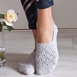 May include: Light grey open-weave socks with a floral design. The socks are worn on feet, with black athletic leggings visible above the ankles. A white rose in a glass vase is in the background.