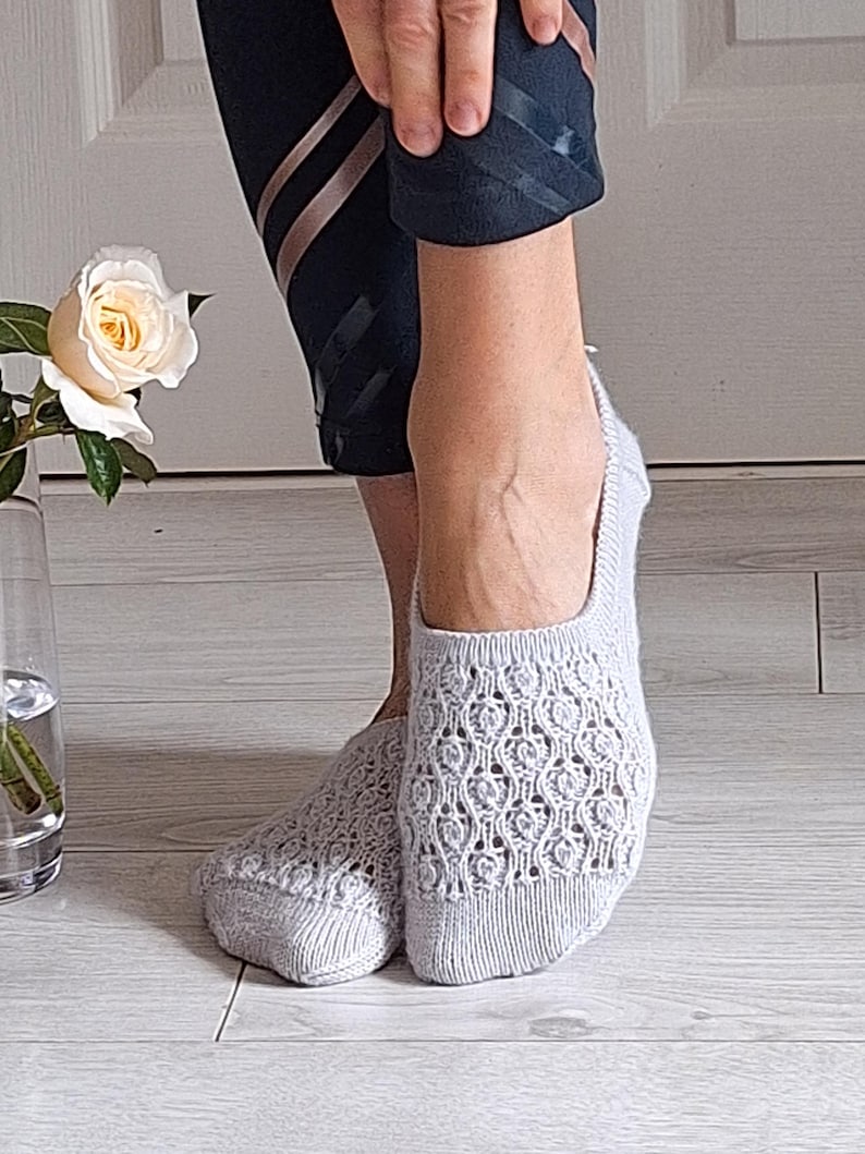 May include: Light grey open-weave socks with a floral design. The socks are worn on feet, with black athletic leggings visible above the ankles. A white rose in a glass vase is in the background.