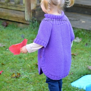 May include: A young girl wearing a purple knitted poncho with a high neck and a textured pattern. She is holding a red leaf in her hand and is standing on a grassy lawn.