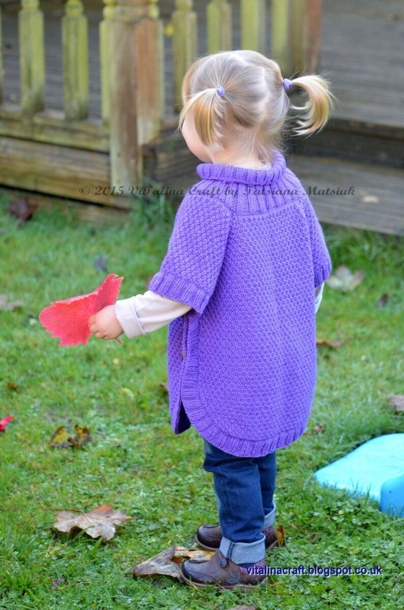 May include: A young girl wearing a purple knitted poncho with a high neck and a textured pattern. She is holding a red leaf in her hand and is standing on a grassy lawn.