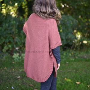 K&ouml;nnte beinhalten: Eine Frau tr&auml;gt einen staubigen rosafarbenen Strickponcho mit geripptem Saum. Der Poncho ist in einem strukturierten Stichmuster gestrickt. Die Frau tr&auml;gt dunkelblaue Jeans und ein graues Langarmshirt.