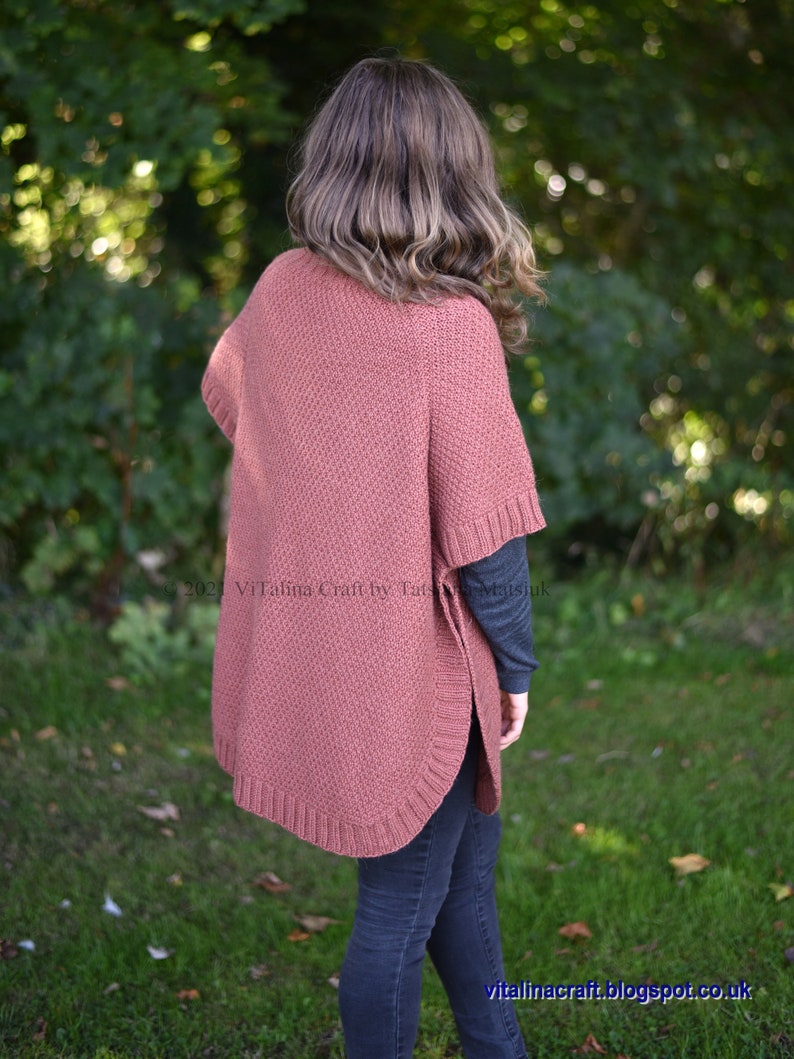 K&ouml;nnte beinhalten: Eine Frau tr&auml;gt einen staubigen rosafarbenen Strickponcho mit geripptem Saum. Der Poncho ist in einem strukturierten Stichmuster gestrickt. Die Frau tr&auml;gt dunkelblaue Jeans und ein graues Langarmshirt.