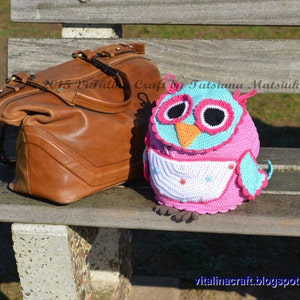 May include: A crocheted owl toy with pink, white, and blue colors. The owl is sitting on a wooden bench with a brown leather bag.
