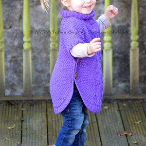 May include: A young girl wearing a purple knitted poncho with a button closure and blue jeans. She is standing on a wooden deck with fall leaves scattered around her.