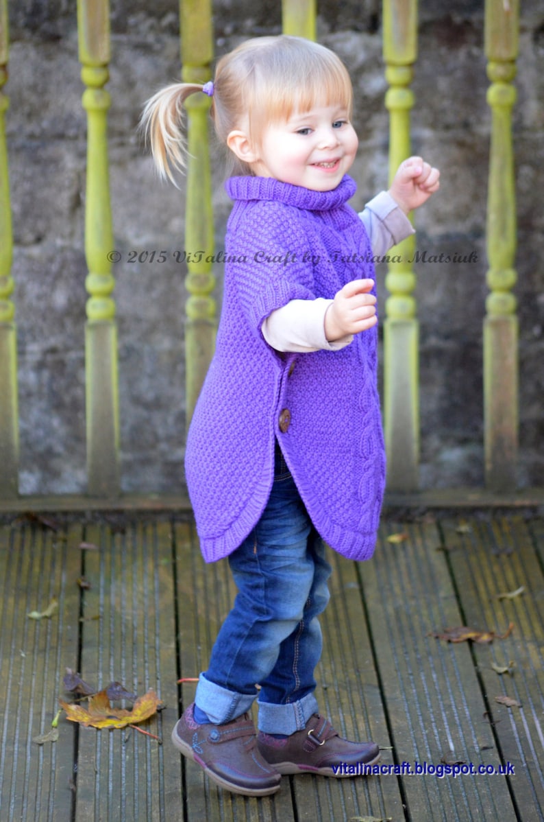 May include: A young girl wearing a purple knitted poncho with a button closure and blue jeans. She is standing on a wooden deck with fall leaves scattered around her.