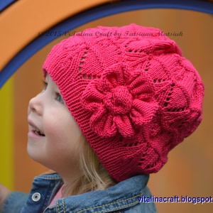 May include: A young girl wearing a pink knitted hat with a flower design. The hat has a lacy pattern and is made with a soft yarn. The girl is looking up and smiling.