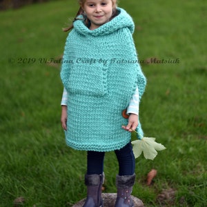 May include: A young girl wearing a turquoise knitted hooded poncho with a wooden button. She is standing on a tree stump in a grassy field. The poncho has a textured knit pattern and is a loose fit. The girl is wearing black leggings and dark blue boots.