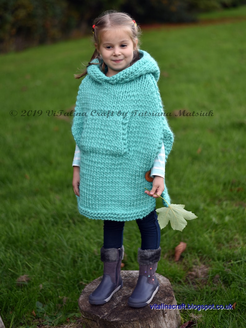May include: A young girl wearing a turquoise knitted hooded poncho with a wooden button. She is standing on a tree stump in a grassy field. The poncho has a textured knit pattern and is a loose fit. The girl is wearing black leggings and dark blue boots.
