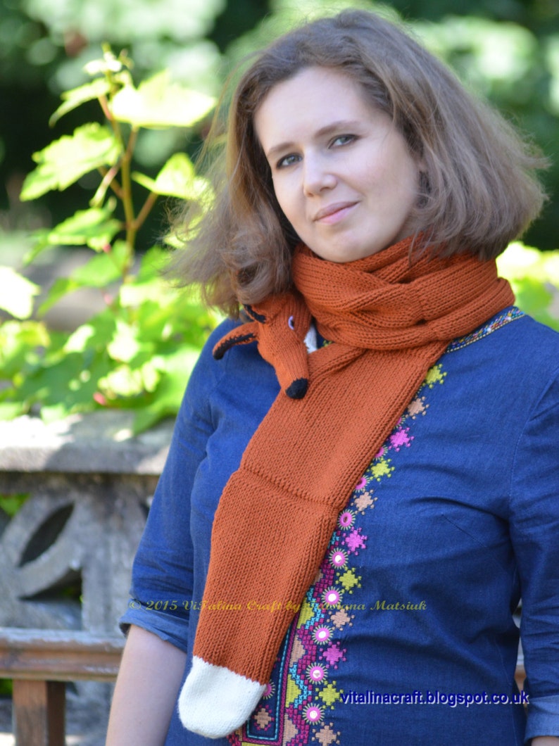 May include: A woman wearing a blue denim shirt with colorful embroidery and a knitted fox scarf. The scarf is orange with white accents and has a fox head at the end.