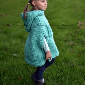May include: A young girl wearing a turquoise knitted hooded poncho with a white and black striped shirt underneath. She is standing in a grassy field.