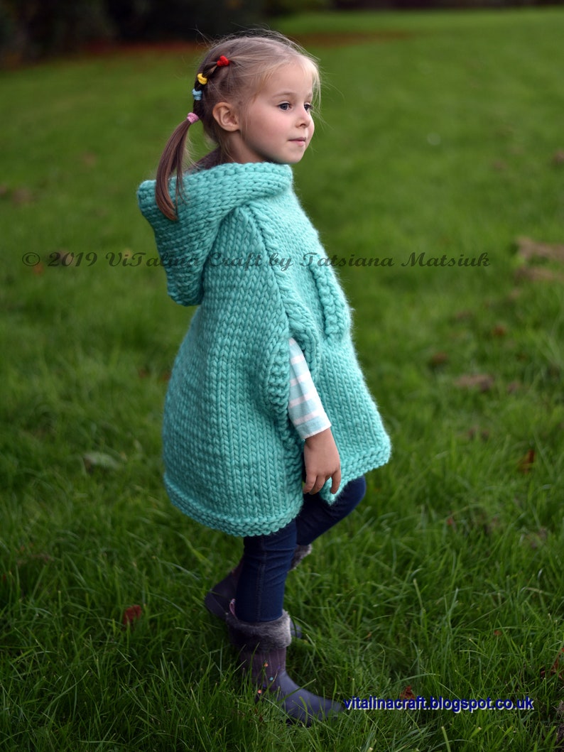 May include: A young girl wearing a turquoise knitted hooded poncho with a white and black striped shirt underneath. She is standing in a grassy field.