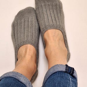 May include: A pair of gray knitted slipper socks. The socks are ribbed and cover the feet. The person is wearing blue jeans with the cuffs rolled up. The background is white.