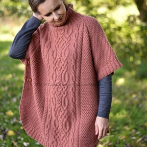 K&ouml;nnte beinhalten: Ein lachsfarbener Strickponcho mit Zopfmuster und hohem Kragen. Der Poncho hat einen Knopfverschluss an der Vorderseite.