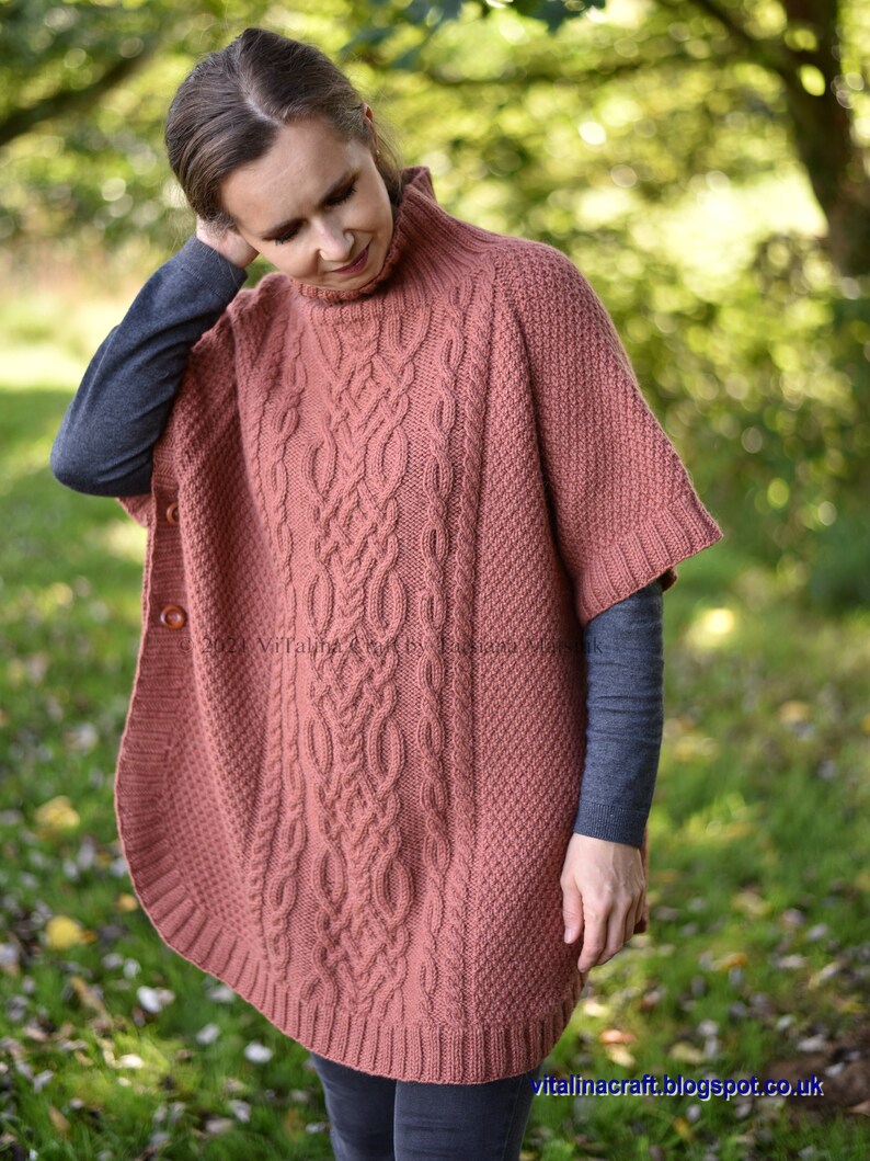 K&ouml;nnte beinhalten: Ein lachsfarbener Strickponcho mit Zopfmuster und hohem Kragen. Der Poncho hat einen Knopfverschluss an der Vorderseite.