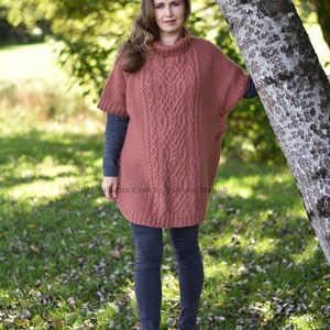 K&ouml;nnte beinhalten: Eine Frau tr&auml;gt einen rostigen Strickponcho mit Rollkragen und einem Knopfdetail. Sie steht in einem grasbewachsenen Bereich mit Herbstlaub auf dem Boden.