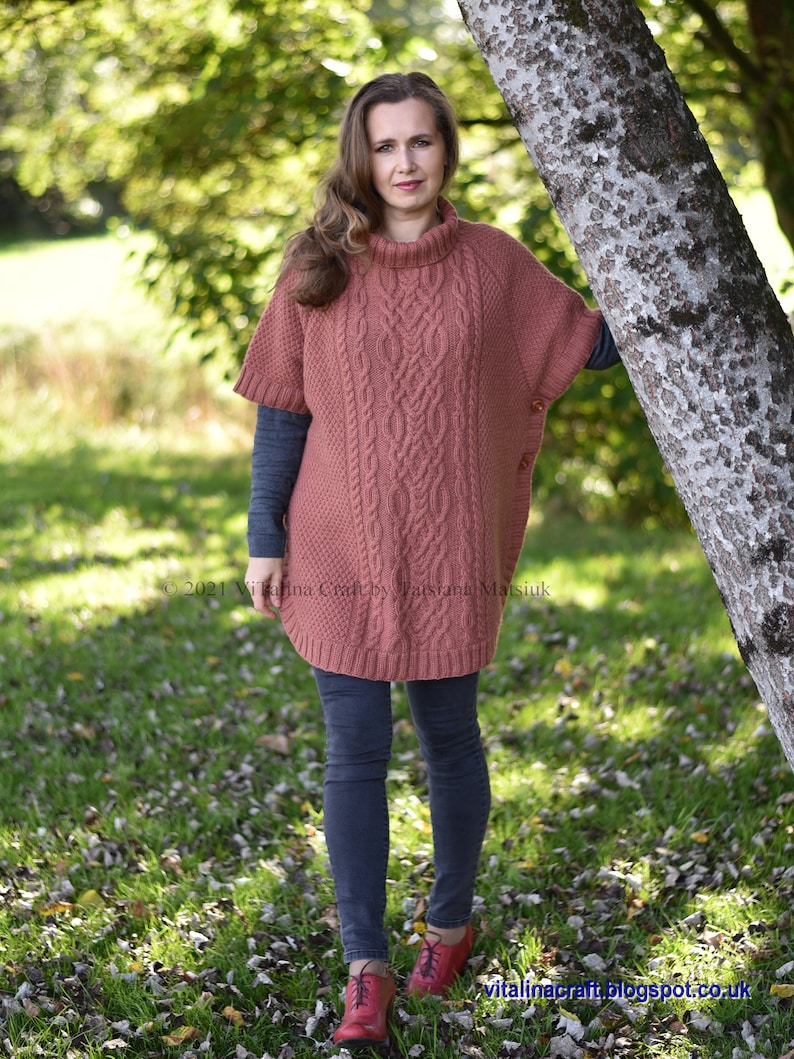 K&ouml;nnte beinhalten: Eine Frau tr&auml;gt einen rostigen Strickponcho mit Rollkragen und einem Knopfdetail. Sie steht in einem grasbewachsenen Bereich mit Herbstlaub auf dem Boden.