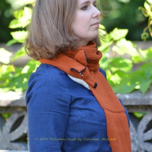 May include: A woman wearing a blue denim shirt with a long, orange and white knitted scarf with a fox face design. The scarf is tied around her neck and hangs down her back. The text "2015 Vitalina Craft by Tatsiana Matsuk" is visible on the scarf.