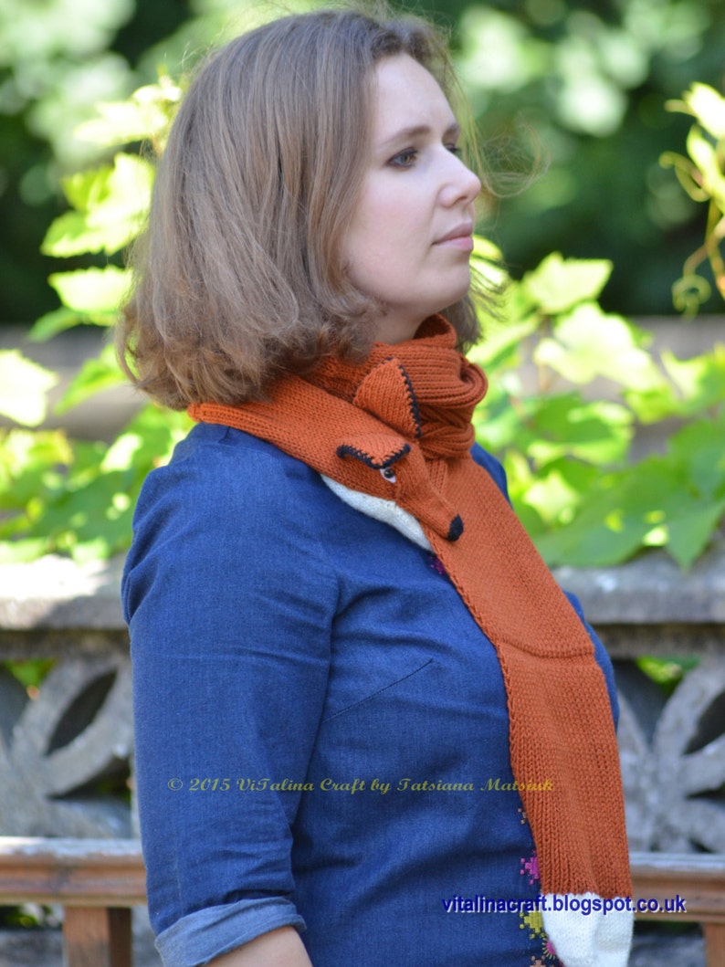 May include: A woman wearing a blue denim shirt with a long, orange and white knitted scarf with a fox face design. The scarf is tied around her neck and hangs down her back. The text "2015 Vitalina Craft by Tatsiana Matsuk" is visible on the scarf.