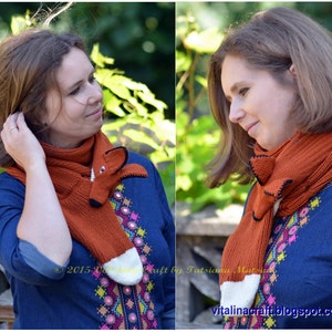 May include: A woman is wearing a knitted fox scarf in orange and white. The scarf has a fox head with black eyes and a black nose. The woman is wearing a blue shirt with colorful embroidery.
