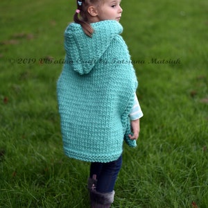 May include: A young girl wearing a teal knitted hooded poncho with a long, loose fit. The poncho is made with a simple knit stitch and has a large hood. The girl is standing in a grassy field.