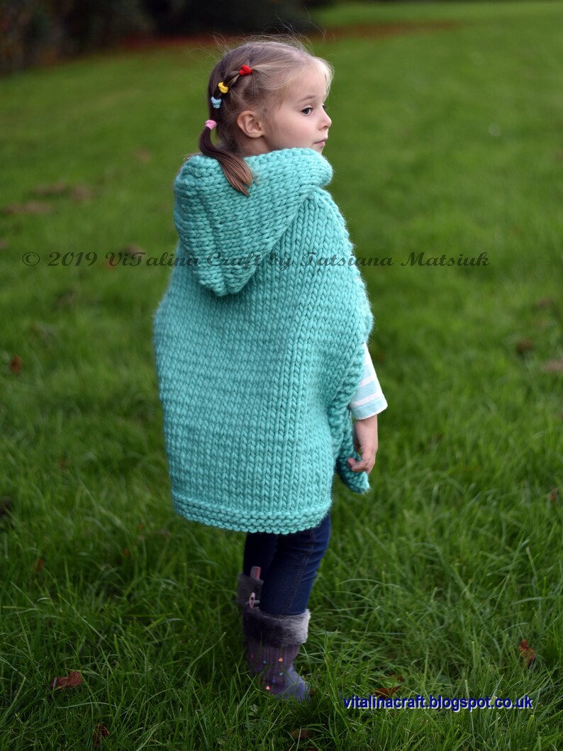May include: A young girl wearing a teal knitted hooded poncho with a long, loose fit. The poncho is made with a simple knit stitch and has a large hood. The girl is standing in a grassy field.