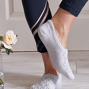 May include: Pale grey knitted no-show socks with an intricate openwork design. The socks are worn with black athletic leggings featuring rose gold stripes. A white rose is in a glass vase.
